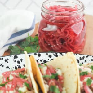 Jar of pickled red onions on cutting board sitting behind a platter of tacos.