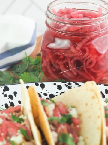 Jar of pickled red onions on cutting board sitting behind a platter of tacos.