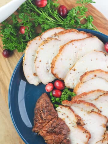 Sliced smoked boneless turkey breast on blue plate with parsley and cranberries on cutting board.